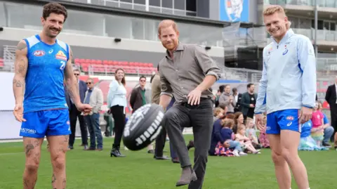 Reuters Prince Harry stands next to two AFL players. He kicks a black ball with white text on it into the foreground.