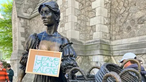A close up of the statue of Molly Malone, but around her neck someone has hung a sign which says 'LEAVE MOLLY mALONE'. 