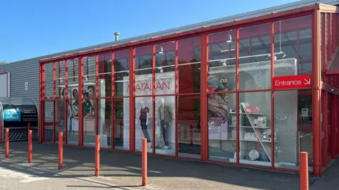 Paul O'Gorman/BBC Outside the Matalan store - a large glass fronted warehouse with red framing and products and advertising images hanging in the windows