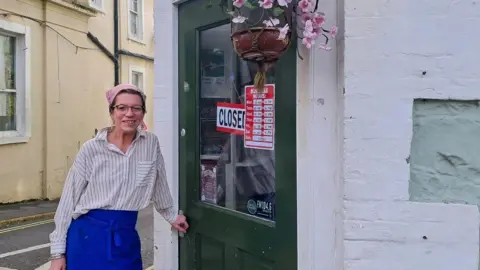 Kirsty Chapman is standing outside her cafe smiling at the camera. She is wearing a blue apron round her waist and has a white and blue stripped shirt on. Her hair is pulled back in a bun and she has glasses on her face.