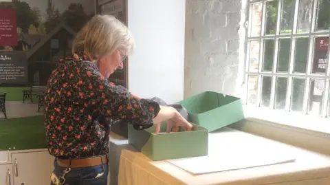 National Trust A woman with grey hair and a blue floral top opening a large pale green box on a white table in front of a window with a number of small panes