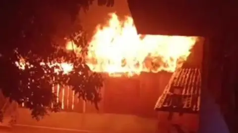 HANDOUT Tall flames are seen behind a fence and gate in a village near the volcano