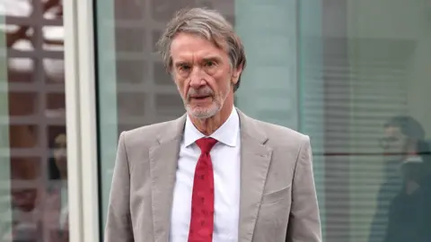 PA Media Sir Jim Ratcliffe looking straight at the camera. He is wearing a beige jacket over a white shirt with a red tie.