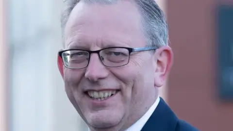 The Conservative Party A man with short grey hair and glasses and wearing a dark blue suit, white shirt and light blue tie is smiling.