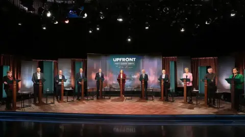 PA Media Ten party leaders stand at podiums during a televised  election debate. The floor and podiums are brown wood and the candidates are looking toward the camera