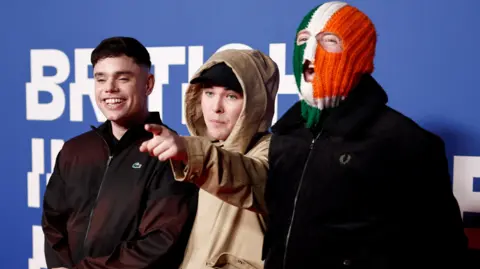 Getty Images The young men pose for the cameras at an awards ceremony. The first man is smiling and wearing a black hoodie, the man in the centre is wearing a brown hoodie, and the man on the right is wearing a green, white and orange ceremony. 