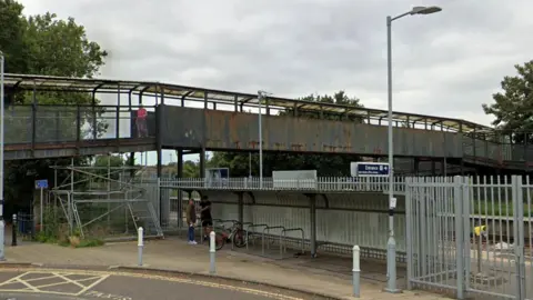 A metal bridge spans a railway. The bridge, which also has a roof, is heavily rusting on its sides.