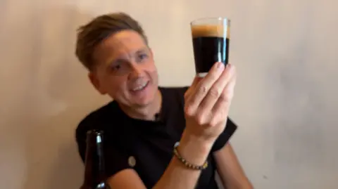 Image shows Ryan holding a beer he has poured into a glass. He is wearing a black top and looking at the beer smiling. The beer is dark in colour and has a foamy top. The beer is in the foreground of the image and Ryan is in the back.