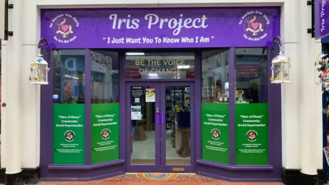 The front of a community social supermarket in Nuneaton. The door and window frames are painted purple and the sign above the front door says 'Iris Project' with the message underneath saying 'I just want you to know who I am'.