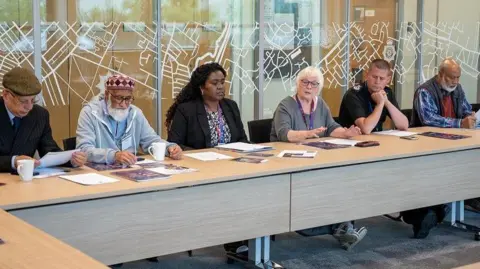 A group of men and woman are sitting behind desks, They include a man wearing a flat cap, a man with a pink and purple Kufi hat, a woman with long dark hair wearing a black jacket, Danielle Stone with white hair and glasses, Ivan Balhatchet in a black police shirt, and a man with a white beard wearing a grey gilet. They are sitting in front of glass panels with white line patterns.