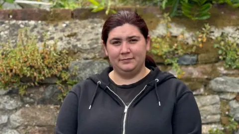 A woman in her mid-20s wearing a black hoodie and a black top. She is stood in front of a stone wall with plants growing out if it