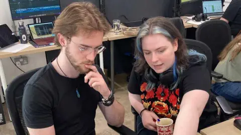 Mousumi Bakshi/BBC Image shows 23 year old Iz Head wearing a printed black and red tee shirt in conversation with a male colleague, wearing a plain black tee shirt