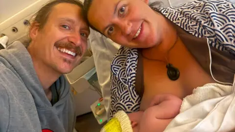 Jenny with her partner smiling at the camera. She is holding her newborn baby who is breastfeeding.