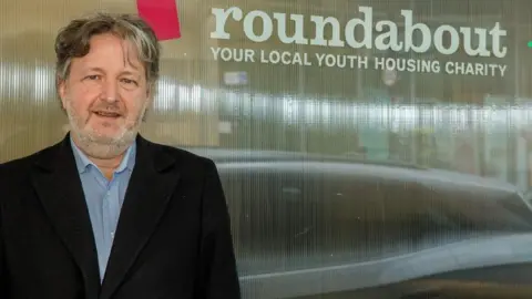 ROUNDABOUT A man with greying hair and a beard in a dark jacket and blue shirt stands next to a window bearing the words 'Roundabout Your Local Youth Housing Charity'.