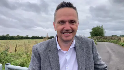 A man stands in the middle of the frame with a neutral expression. He's wearing a grey suit and white shirt. Behind him are fields and a road.
