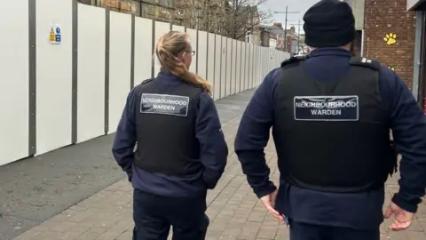 A man and a woman pictured from behind walking down the street. They are both wearing a blue uniform with a black vest that says 'neighbourhood warden'. The woman has long blonde hair in a ponytail and wears glasses. The man wears a black beanie hat and has a beard.