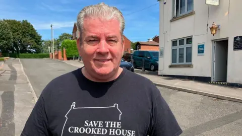 A grey-haired man wearing a black t-shirt is looking directly at the camera. He is standing outside a white-painted pub with cars parked in a car park to the side. The man's t-shirt reads "Save The Crooked House" in white letters.