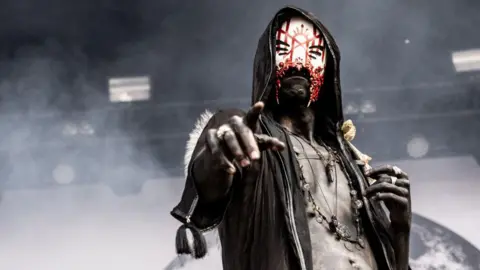 A member of Sleep Token on-stage looks into the camera, one hand with finger pointing at the viewer. He wears a black hooded cloak, leather vest and a white mask with three eyehole slits on each side and an ornate red pattern neatly painted on.