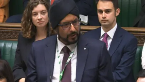 Warinder Juss, wearing a dark suit, stands in the House of Commons, flanked by other MPs sitting behind him
