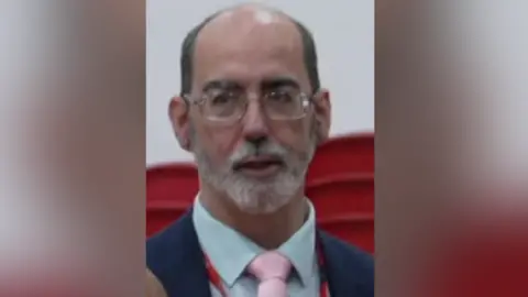 BBC Anthony Felton is looking towards the camera and standing in front of piled up red plastic chairs in front of o a white wall. He is bald on top with close cropped dark hair on the sides. He is wearing glassed and a has a groomed white beard. 