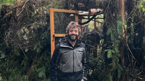 Ed Parr Ferris, from the Devon Wildlife Trust, standing in front of a soft-release enclosure in woodland, which has an open gate