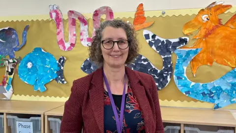 Headteacher Clare Welbourne wearing dark rimmed glasses and a burgundy blazer. She is in front of wall with children's under water fish art on it 