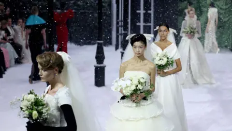 Isabel Infantes/Reuters Four models wearing white wedding dresses stand in a line on a snow-covered stage and snow is falling on them