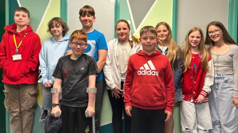 9 students from year 7 and 8 stand smiling for a group photo. They are dressed in non uniform. The wall behind them is covered in geometric shapes which are painted green, blue, grey and yellow.
