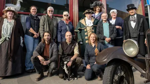 A group of people in Victorian-era outfits stand outside a shop smiling at the camera. There are historic vehicles in the foreground.