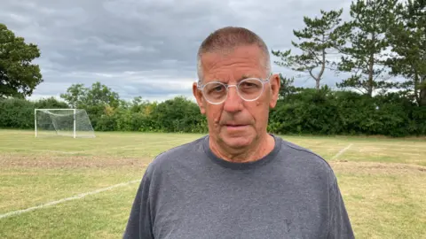 A man stands in a football field, wearing a dark grey t-shirt and clear glasses.