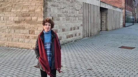 Supplied Carrie Terry (woman, red scarf, blue patterned jumper) on a paved area in front of the stone walls of the Ada Belfield centre.