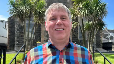 Glen Simpson is smiling as he stands in front of a church building covered slightly by two tall trees. he has short greying hair and is wearing a checked, multi coloured shirt.
