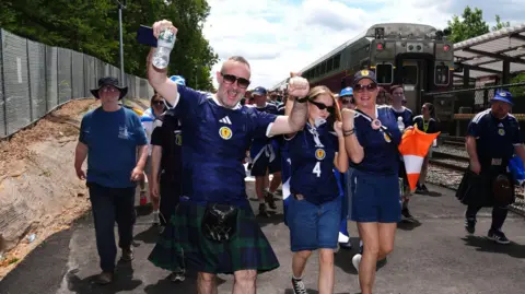 PA Media A group of people wearing Scotland tops alight off a train onto the platform. They pose, some with their hands in the air, while walking towards the camera in summery clothes and some kilts