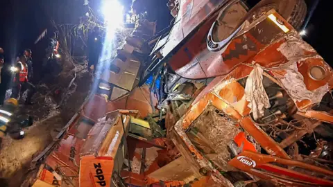 Wreckage of the bus seen in the dark. There is a glaring light in the background from what appears to be a torch. The wreck is on the right. on the left are people in high-vis with torches.