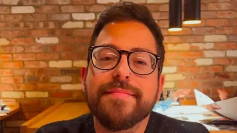 A head and shoulders image of a man wearing glasses and with a nose ring. He is smiling and looking into the camera. 
