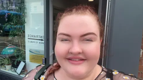 Lauren has brown hair pulled back in a plait and is wearing a mesh black top with flowers on it.