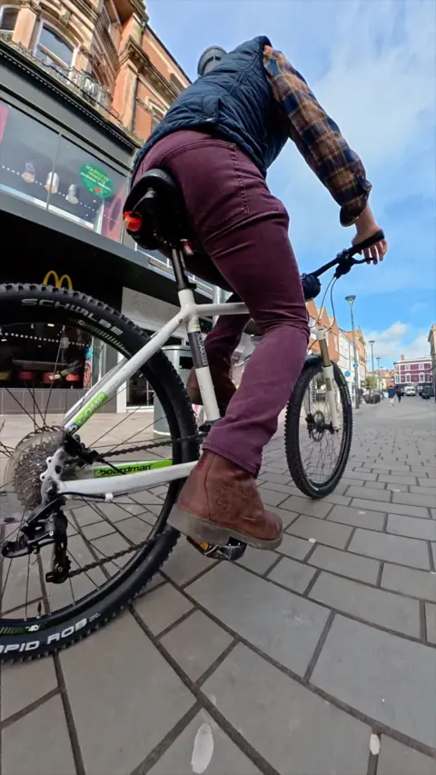The image shows a low‑angle view of a person riding a white mountain bike through Derby city centre. The shot focuses on the bike’s rear wheel and the rider’s legs as they pedal along a paved pedestrian street. Shopfronts, including a McDonald’s, line the background under a bright sky.
