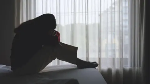 Getty Images A stock image of a woman sitting and crying on a bed in bedroom. She is in darkness. Some light is coming in from the sheer curtain and some trees and buildings can be seen outside through it.