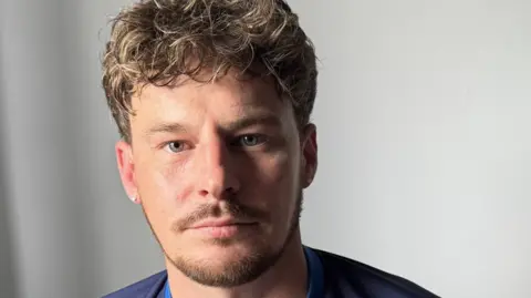 A man with curly brown hair and a thin beard in front of a grey background