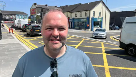 Barney Regan with the box junction and cars behind him. He has short grey-ish hair, wearing a light blue t-shirt with sunglasses hanging from the neck.