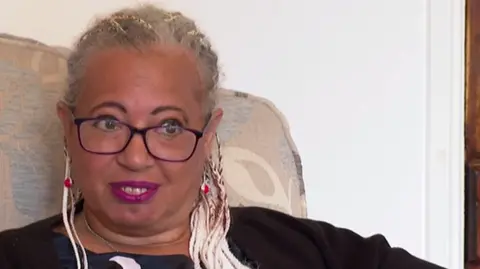A woman with blonde braided hair sits in a chair with a floral beige pattern on it. She has thin glasses on and purple lipstick. 