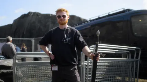 Courtesy of HBO and Creative Wales Aaron on set in sunglasses and black top holding a sword, with temporary metal fencing behind him and a truck