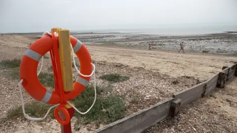 PA Images A bright orange life ring hanging on a pillar. The life ring has two white stripes and has been propped up on the beach. Shingle, sand and the sea are in the background.