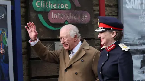The King, wearing a brown wool double-breasted coat, waves to the crowds as he stands under a sign that says Clitheroe Interchange. The Lord-Lieutenant of Lancashire, Mrs Amanda Parker, smiles next to him in a navy uniform with red and gold trims