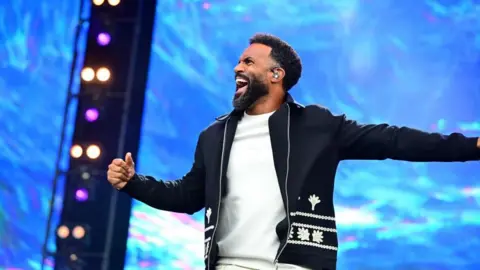 Craig David in a white t shirt and dark jacket, he is mid-performance and has a joyful look on his face