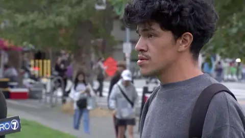 A man with a grey t-shirt and black curly hair speaks to camera.