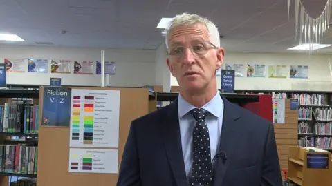 A shot of Chris Prosser from the torso up. He is dressed smartly in a suit and tie, and has grey hair and glasses on. Behind him is a school library.