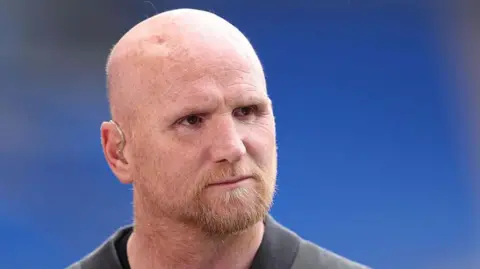 Getty Images John Hartson has a bald head and a ginger beard. He has an earpiece in his ear. He is looking off camera and there is a blue background