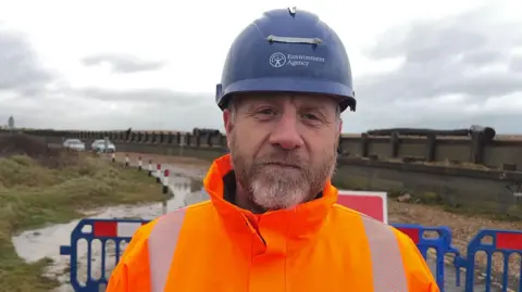 BBC/George Carden A man with an orange high vis and a blue hard hat looks at the camera. He is standing in front of a fenced off shingle beach