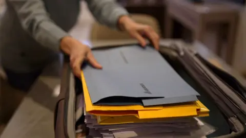 Brijesh Patel / BBC A person is organizing a stack of documents inside a suitcase. The documents include blue and yellow folders, with the top blue folder labelled 'CONFIDENTIAL.' The person's hands are visible, arranging the folders neatly. The background shows a blurred indoor setting with furniture.
